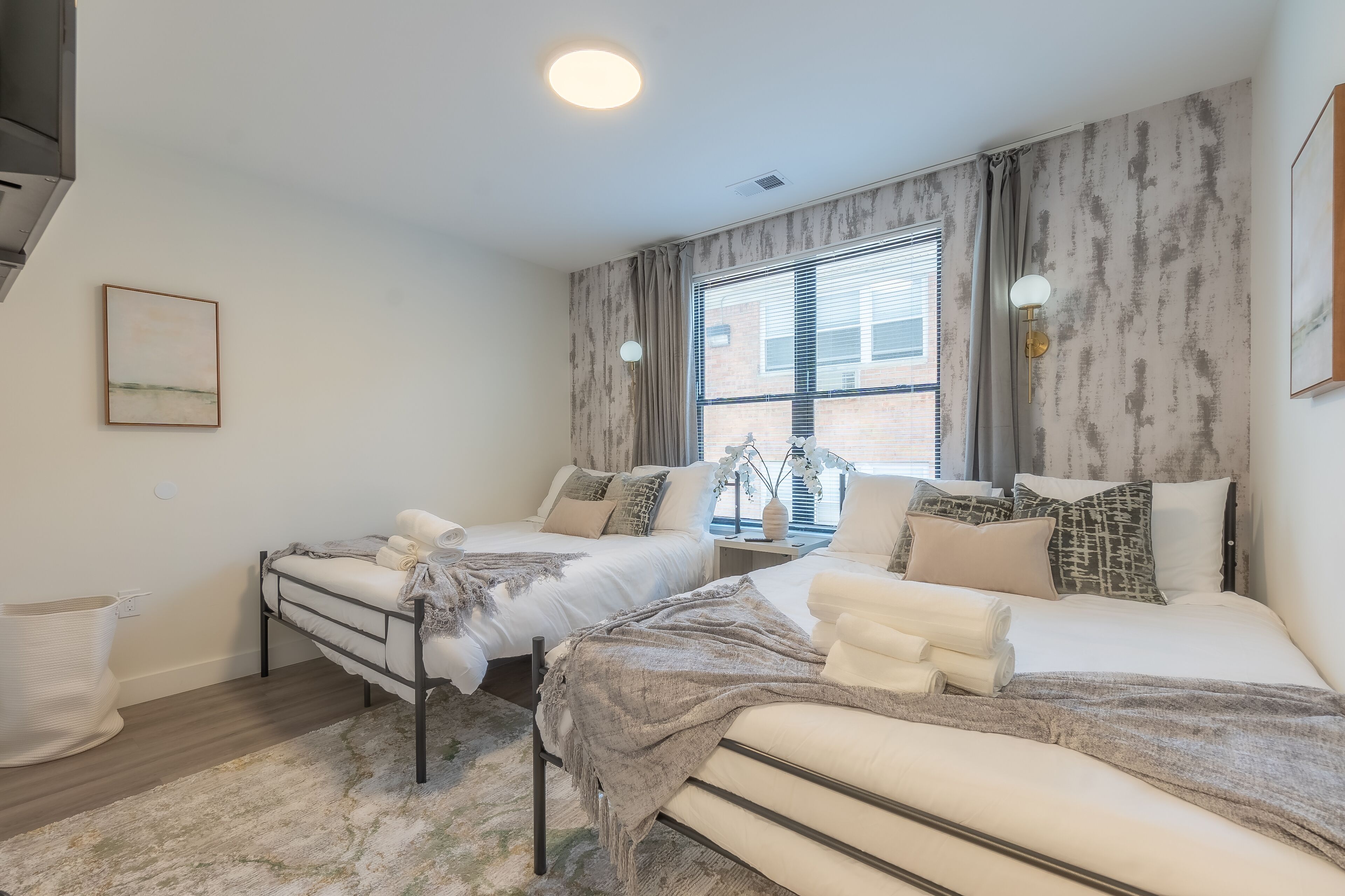 A modern bedroom with two beds, a large window with curtains, a bedside table with a lamp, and a wall-mounted artwork.