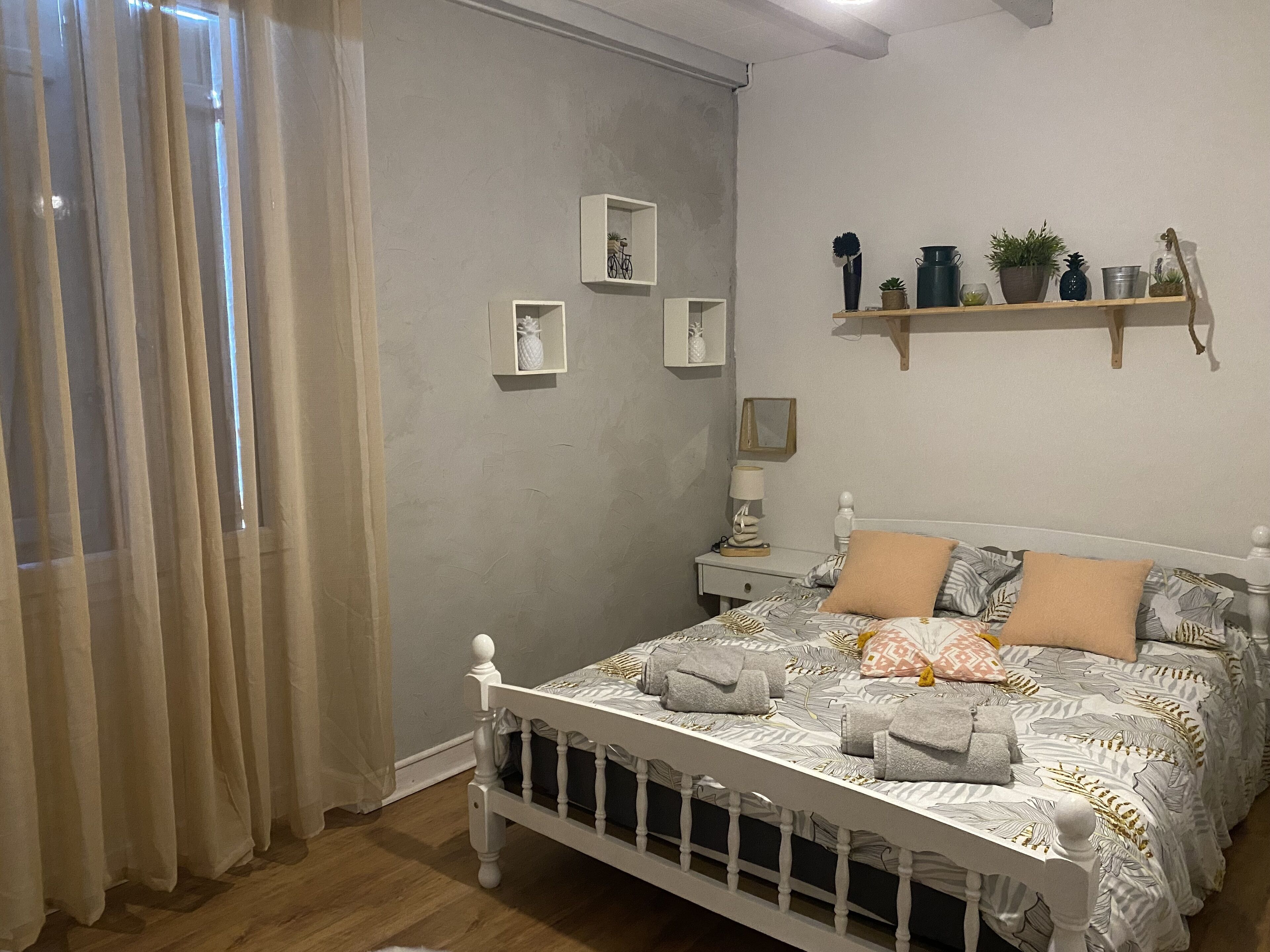 A bedroom with a bed, a nightstand, a shelf with plants, and a window with curtains.