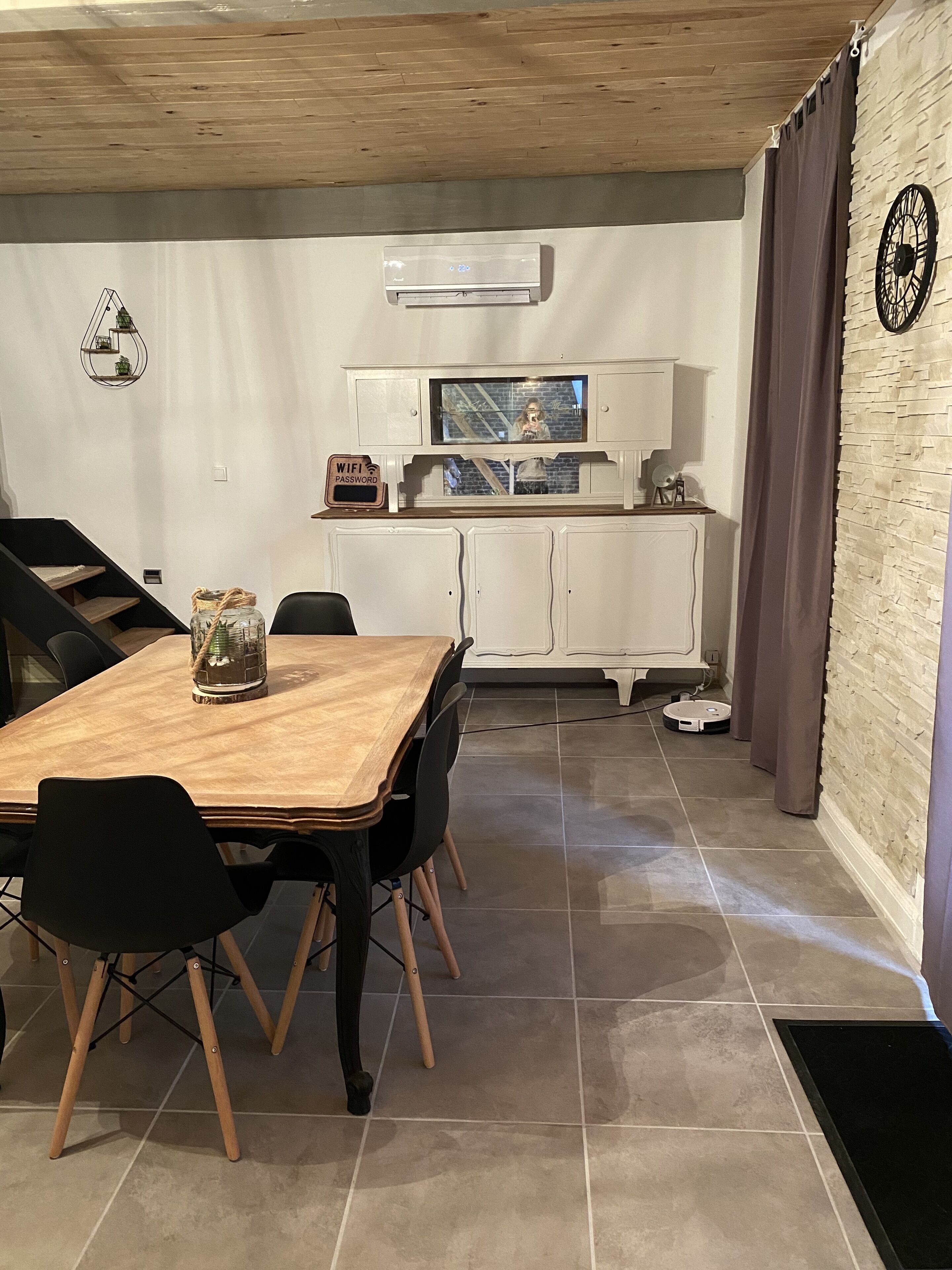A dining area with a wooden table and chairs, a kitchen with white cabinets and a built-in oven, and a brick wall with a clock.