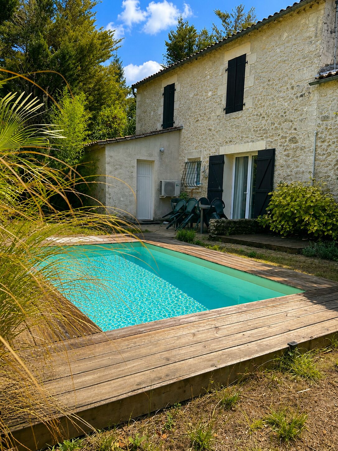 Una piscina con muelle de madera, rodeada por un edificio de piedra con persianas y abundante vegetación.
