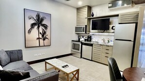 Un salon moderne avec un canapé gris, une table basse en bois et un coin repas comprenant une table et des chaises. On aperçoit une cuisine équipée d’un four à micro-ondes, d’une cuisinière et d’un réfrigérateur.