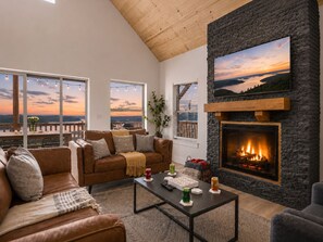 A living room with a stone fireplace, a large TV, and a view of the sunset through large windows. - Hot Tub, Pool, Lake Views, King Beds - Sleeps 18 (Branson)