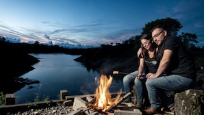 Un couple est assis près d’un feu de camp, face à une rivière paisible, au crépuscule.