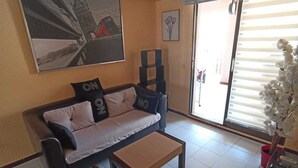 Una sala de estar con sofá, mesa de centro y una mesa de comedor con sillas. Hay un ventanal con persianas, una planta en maceta y un aire acondicionado instalado en la pared.
