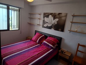 Une chambre à coucher avec un lit, des tables de chevet, une commode, une fenêtre avec des barreaux et des étagères fixées au mur.