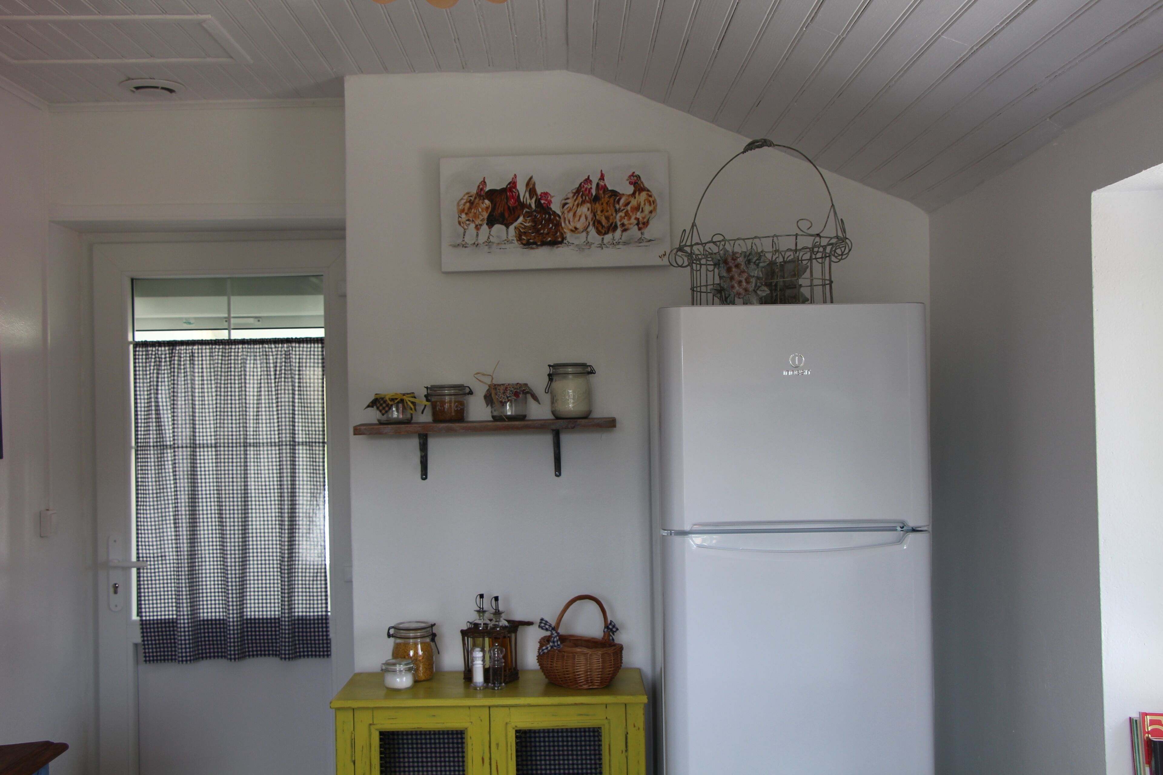 A kitchen with a white refrigerator, a yellow cabinet, and a window with checkered curtains.
