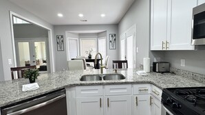 Una cocina moderna con gabinetes blancos, encimeras de granito, electrodomésticos de acero inoxidable y un comedor visible a través de un vano abierto.