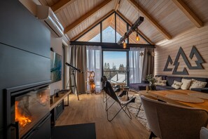 A cozy cabin interior with a fireplace, wooden walls, a large window, and a mountain view.