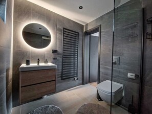 A modern bathroom with a round mirror, a wooden vanity, a toilet, and a glass shower enclosure. - Luxury Chalet with Hot Tube & Sauna (Liptovský Mikuláš)