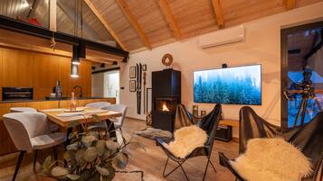 Ein modernes Wohnzimmer mit Kamin, Essbereich und einem großen Fernseher, der eine Waldszene zeigt.
