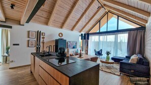Eine moderne Küche mit einer zentralen Kochinsel, Holzbalken, großen Fenstern und einer gemütlichen Sitzecke mit Sofa und Sesseln.