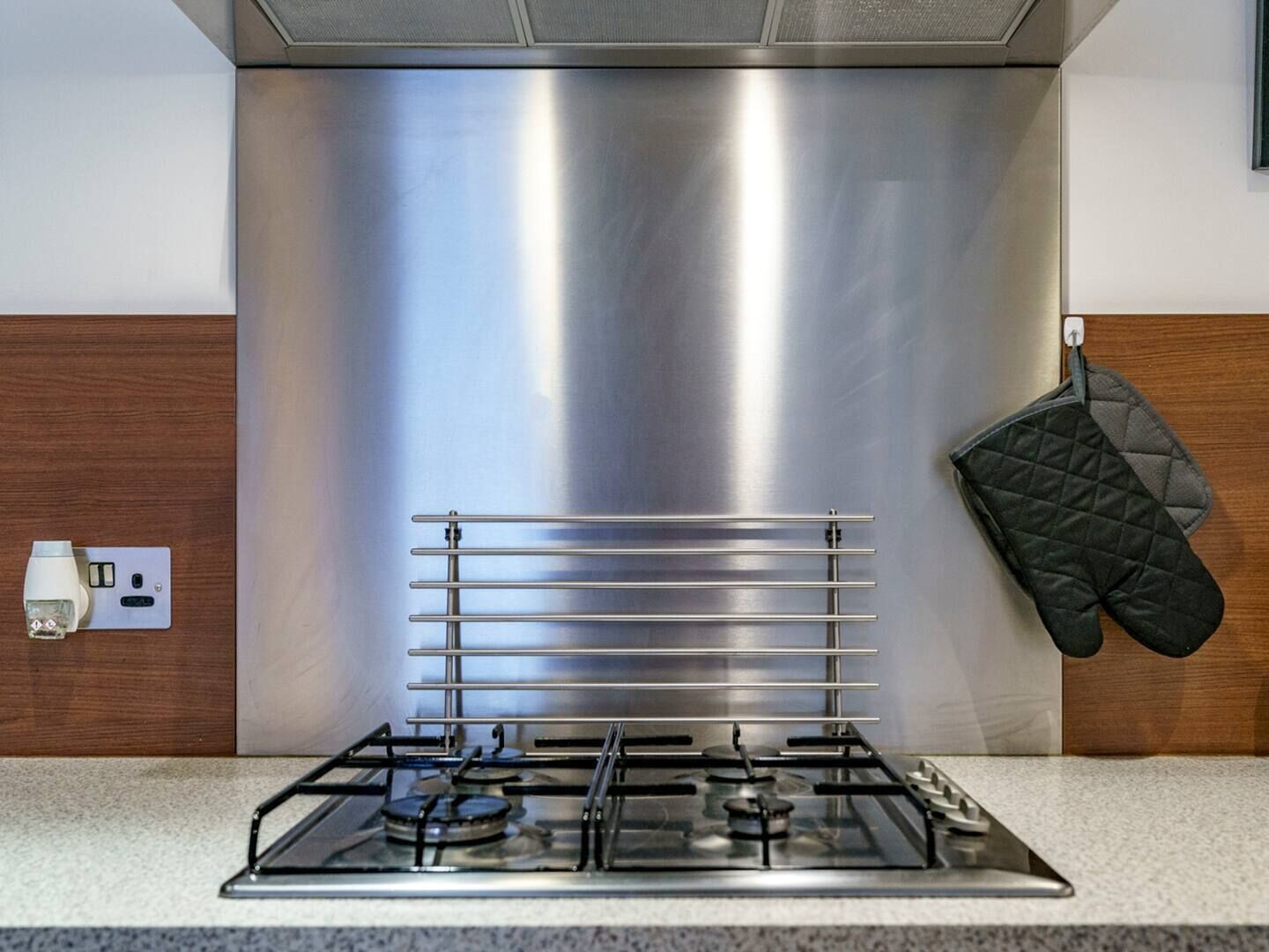 A stainless steel range hood above a gas stove with a wooden backsplash and a black oven mitt hanging on the wall.
