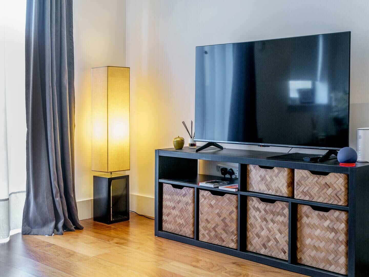 A modern living room with a large flat-screen TV, a floor lamp, and a herringbone-patterned storage unit.