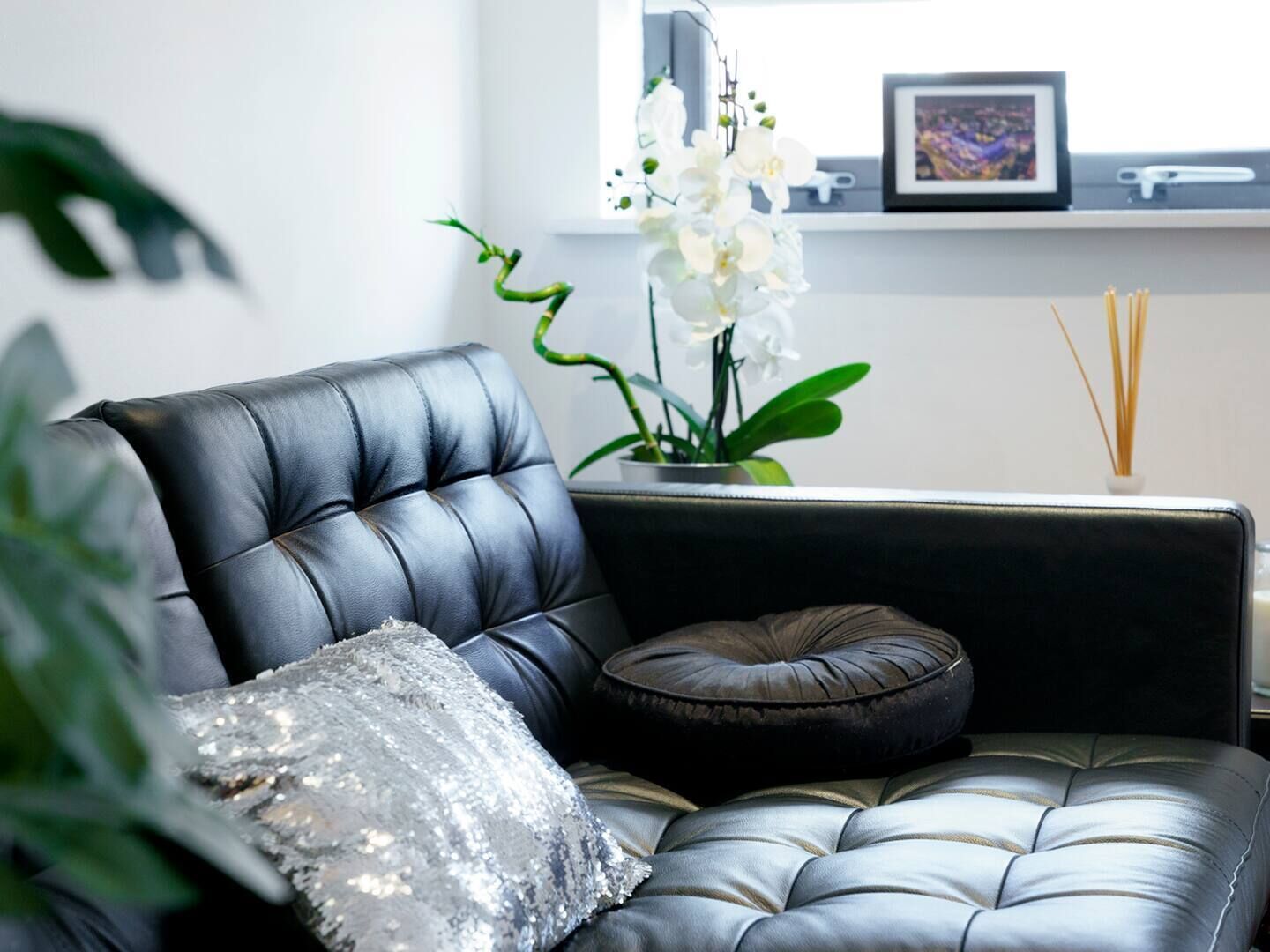 A black leather sofa with a sequined pillow and a dark cushion, a small potted orchid, and a picture frame on a shelf.
