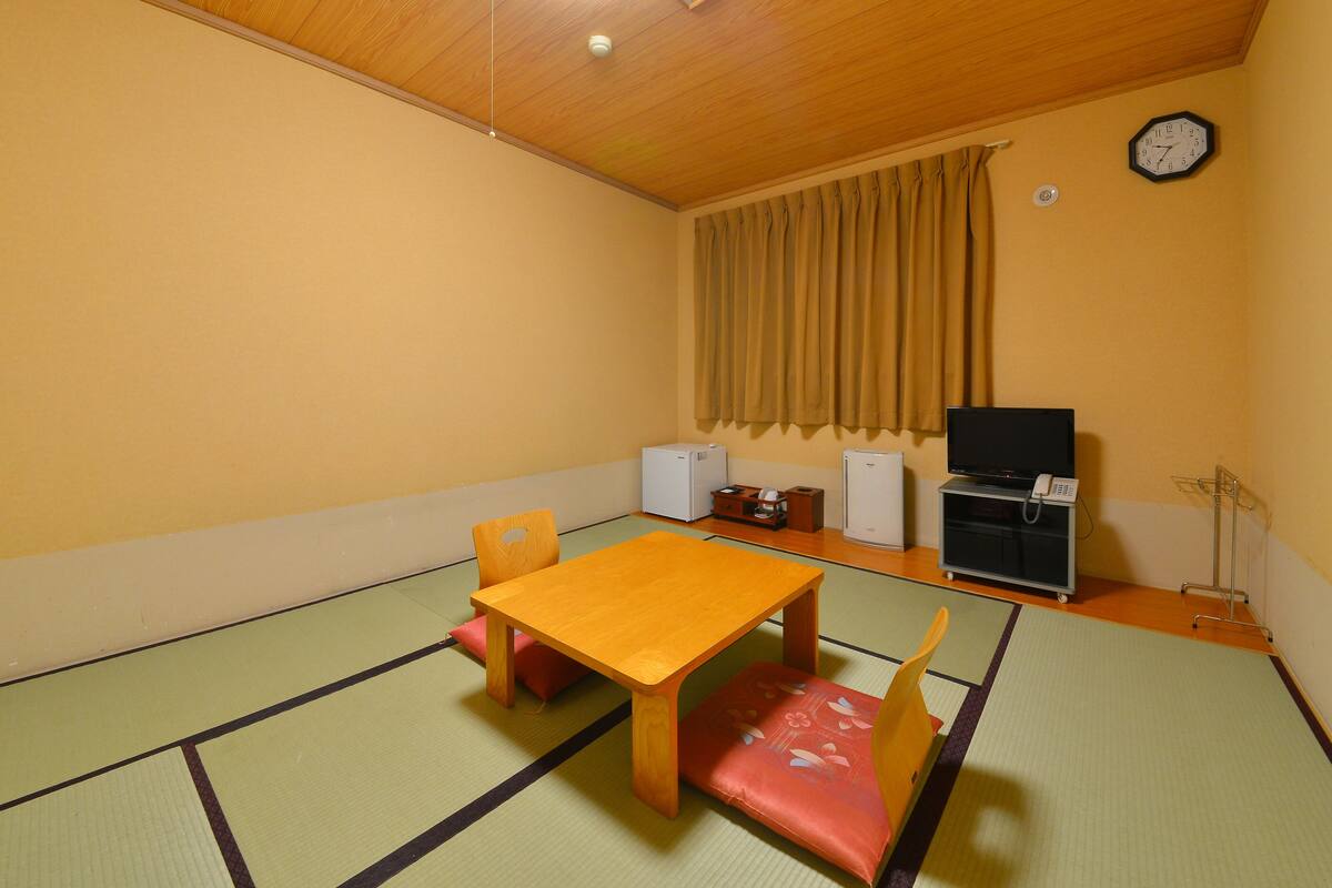 A room with tatami flooring, a wooden table, and chairs. There is a clock on the wall and a television on a stand.