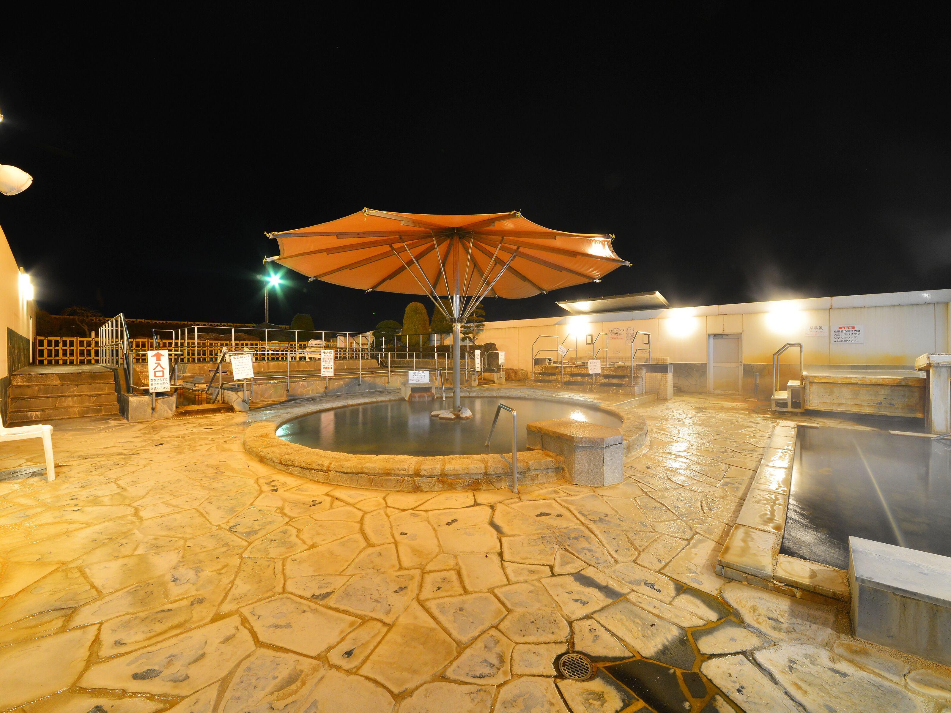 A stone-floored outdoor pool area with a central hot tub, surrounded by steps and a large umbrella.
