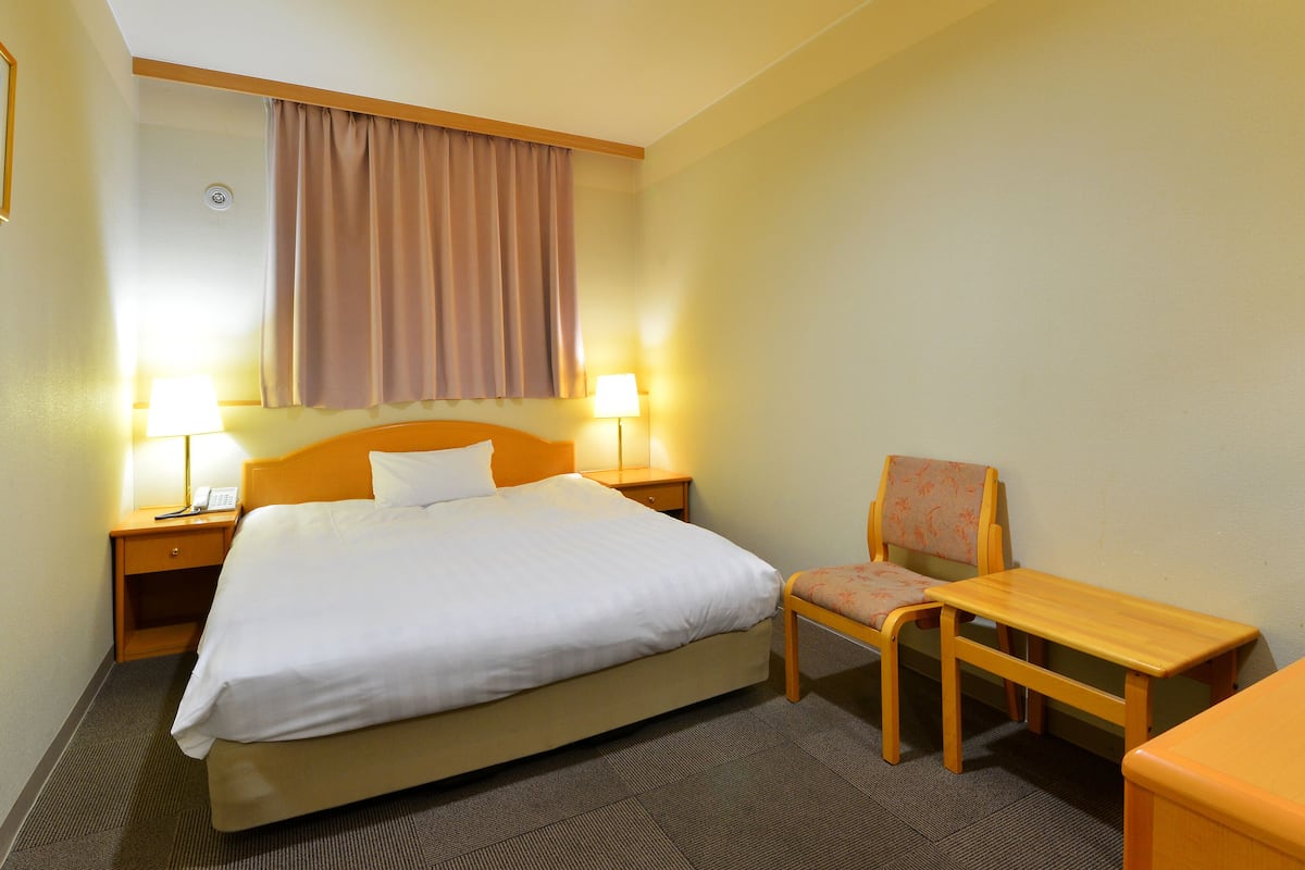 A hotel room with a bed, two bedside lamps, a chair, a small table, and a window with curtains.