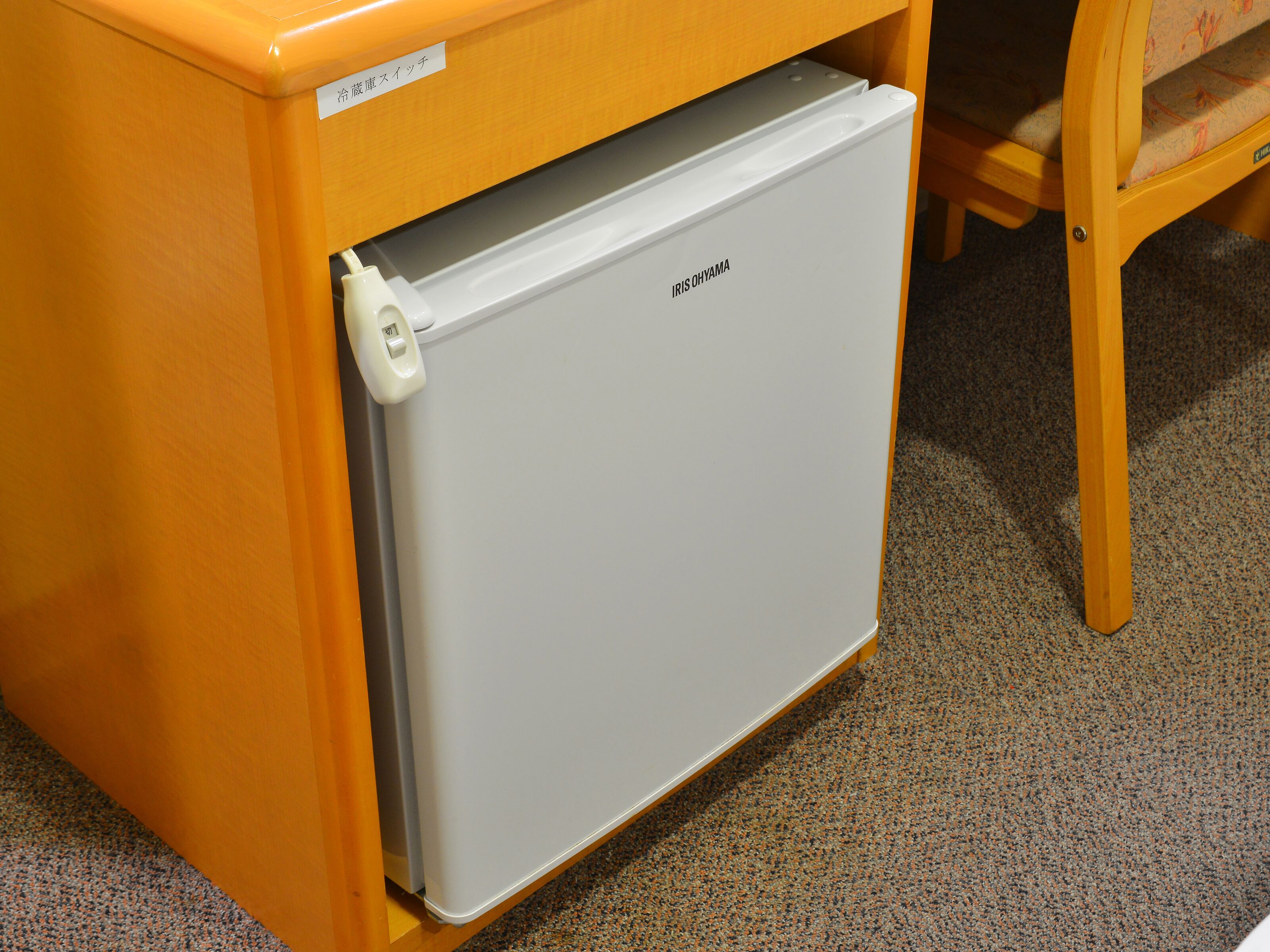 A compact, white refrigerator with a control panel on the top, placed under a wooden table with a cushion.