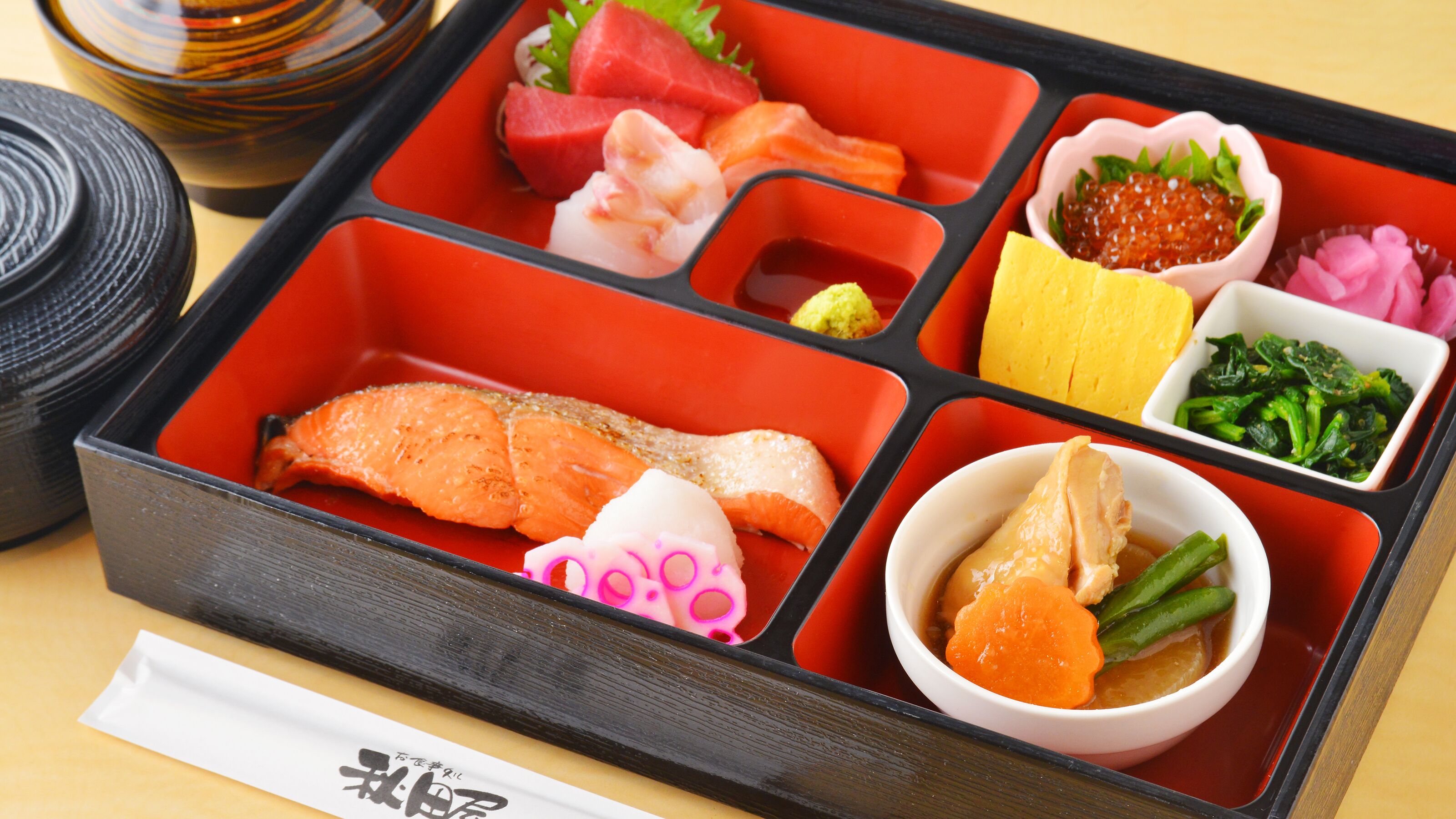 A bento box with various food items including fish, vegetables, and a small bowl of soup.