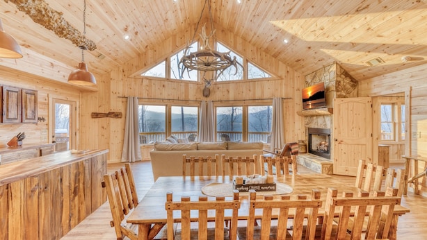 L’intérieur d’une cabane en rondins comprend un coin repas, des meubles en bois, une cheminée et de grandes fenêtres.
