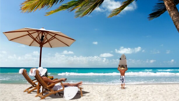 A beach scene with a wooden umbrella, two lounge chairs, and a person walking with a surfboard.