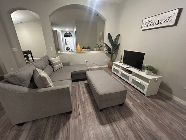 Un salón con sofá modular, reposapiés, mueble para televisor y un cartel en la pared que dice "blessed".
