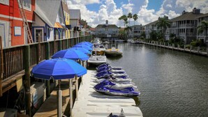 A waterfront area with boats docked along a wooden pier, a row of colorful buildings, and a clear sky.