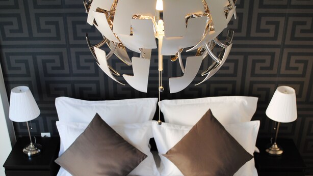 Una cama bien tendida con sábanas blancas y dos almohadones marrones, flanqueada por dos mesitas de noche con lámparas, frente a una pared con diseño geométrico.