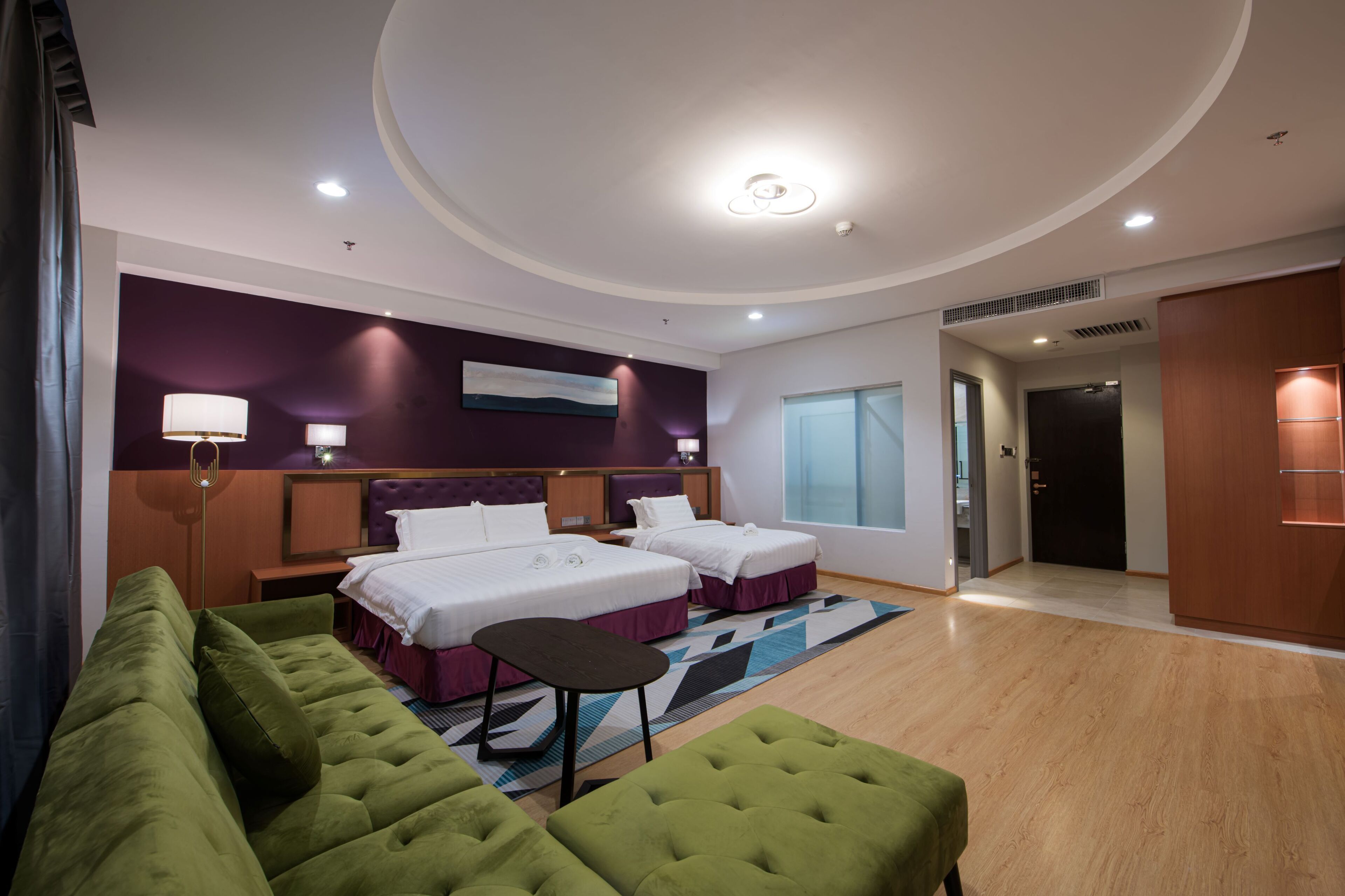 A hotel room with two beds, a green sofa, a small table, and a large window.