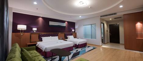 A hotel room with two beds, a green sofa, a small table, and a large window.