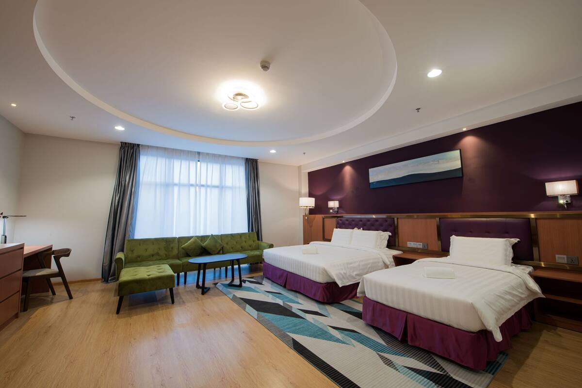 A hotel room with two beds, a green sofa, a small table, and a large window with curtains.
