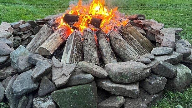 Ein Lagerfeuer in einer steinernen Feuerstelle, umgeben von einem grasbewachsen Feld mit Bäumen und einem Gebäude im Hintergrund.