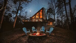 Una cabaña con techo triangular, un hoyo de fuego con sillas y un cielo nocturno lleno de estrellas.