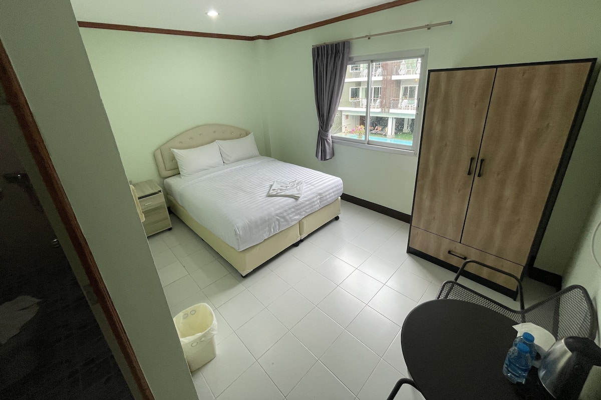 A bedroom with a bed, a wardrobe, a window with curtains, a small table with a chair, and a trash bin.