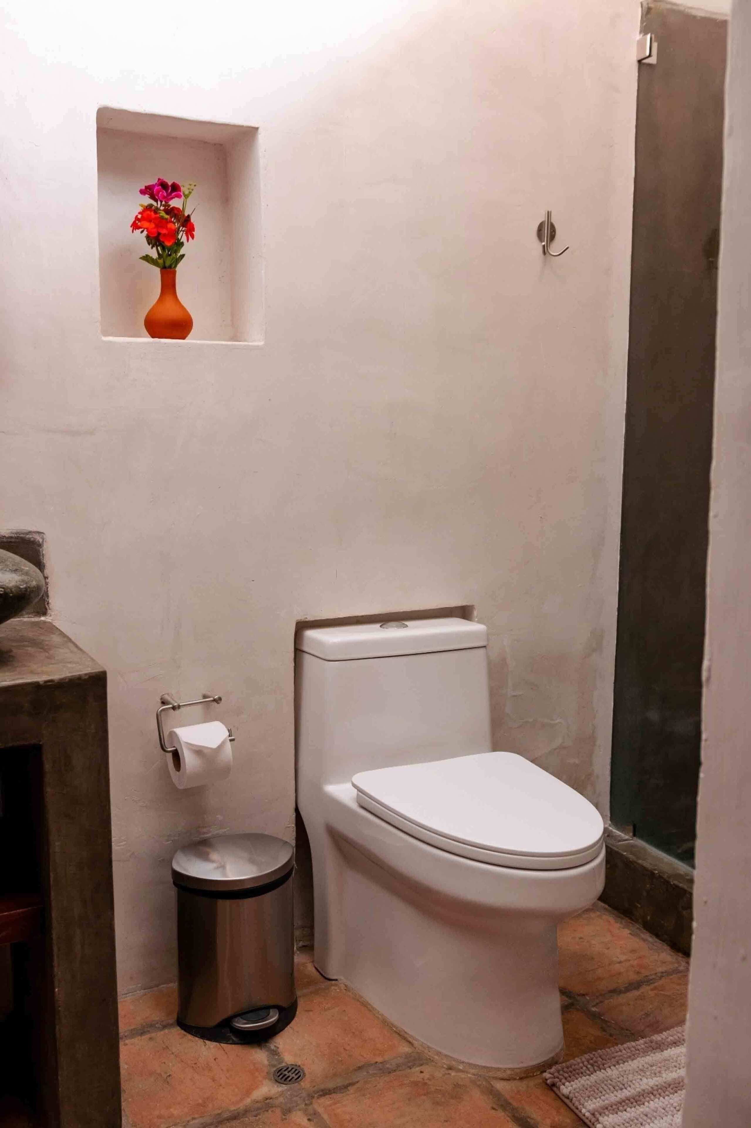 Un baño pequeño con inodoro blanco, un bote de basura y un estante con un jarrón de flores.