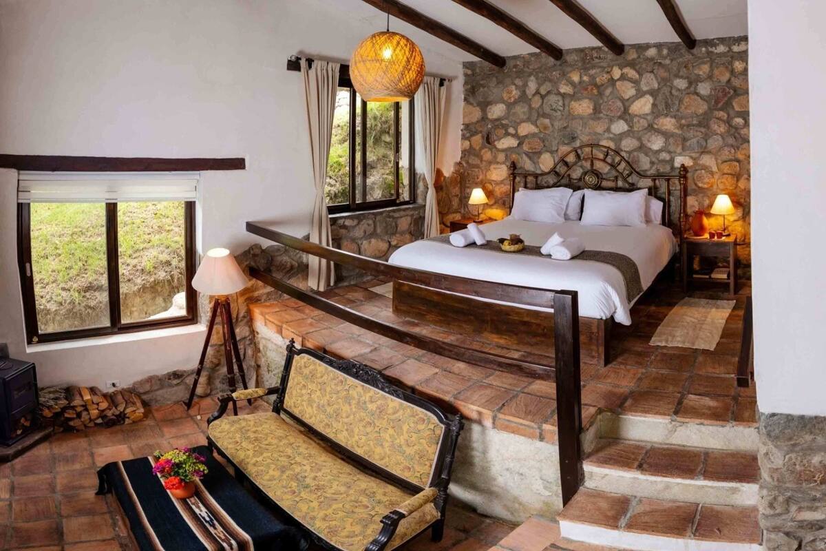 A rustic bedroom with a stone wall, a wooden bed, a bench, and a hanging light fixture.