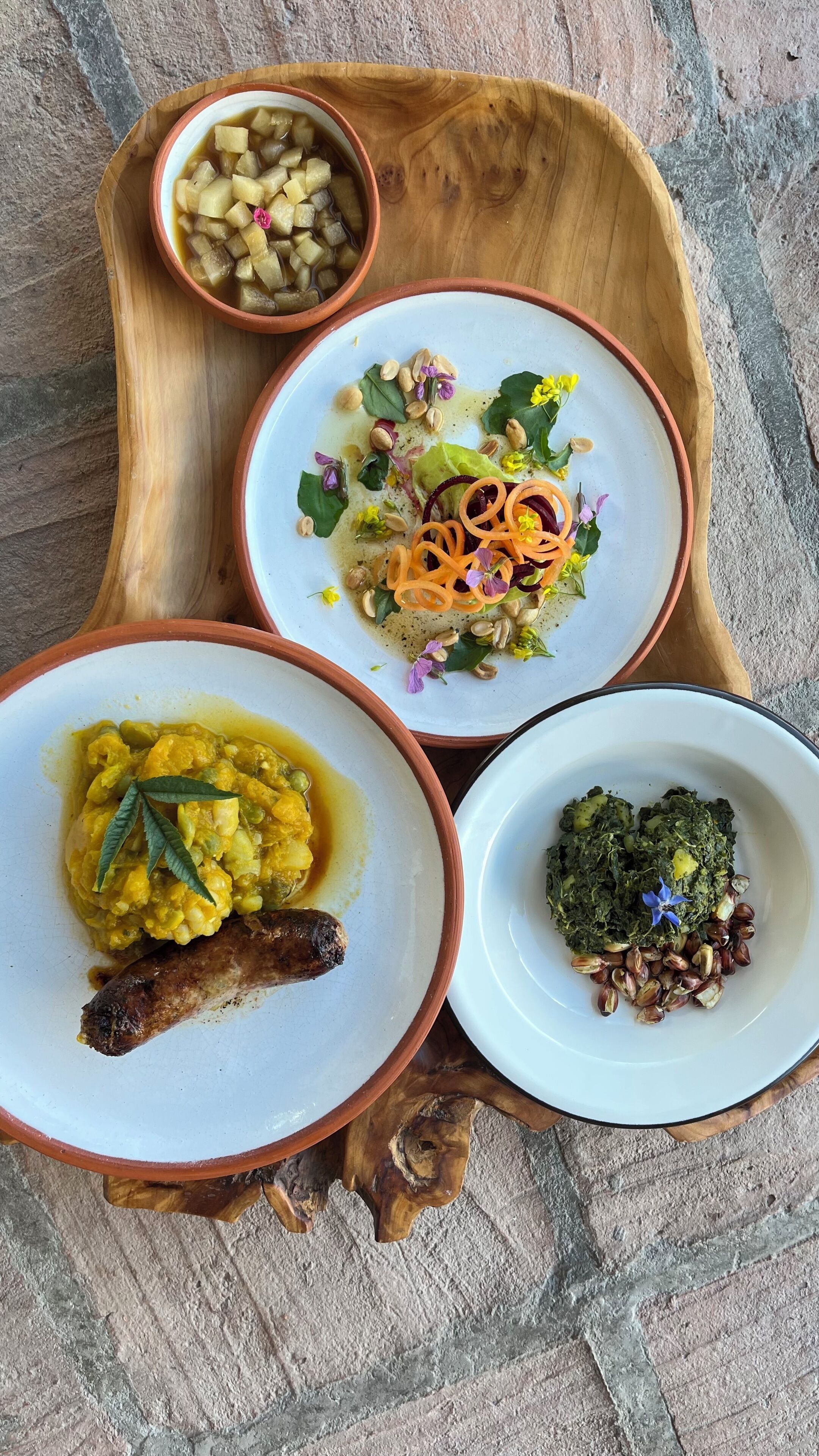 A wooden tray with four dishes: a bowl of diced potatoes, a salad with nuts and greens, a bowl of yellow curry with a sausage, and a dish with greens and nuts.