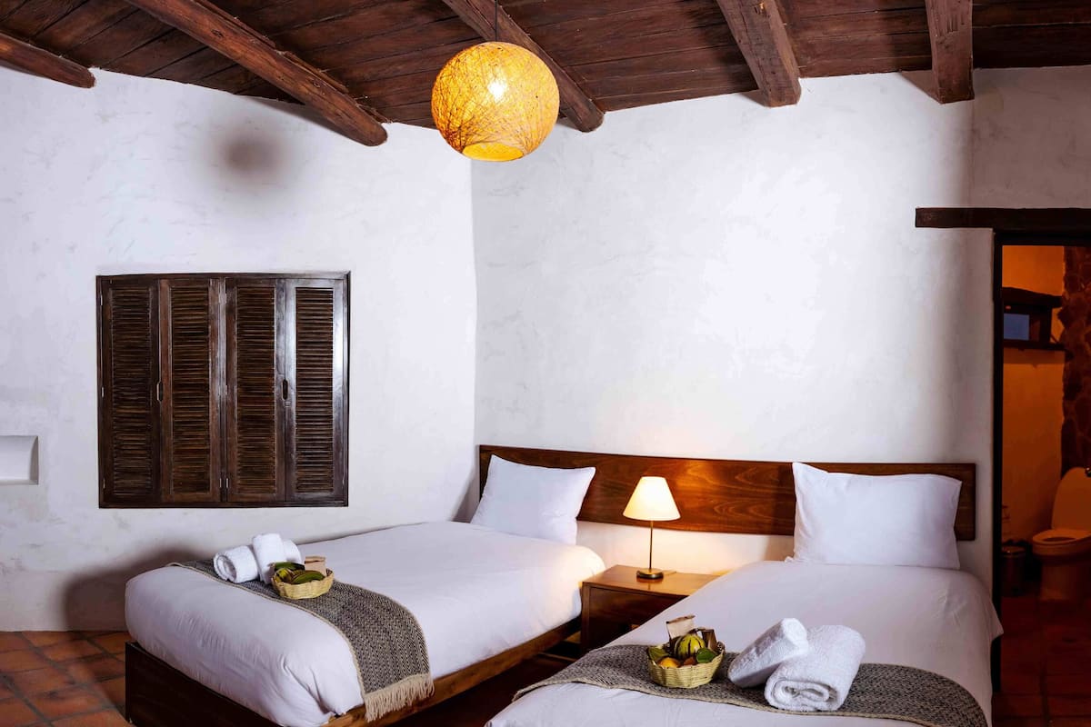 A room with two beds, a wooden ceiling, a hanging light fixture, and a window with shutters.
