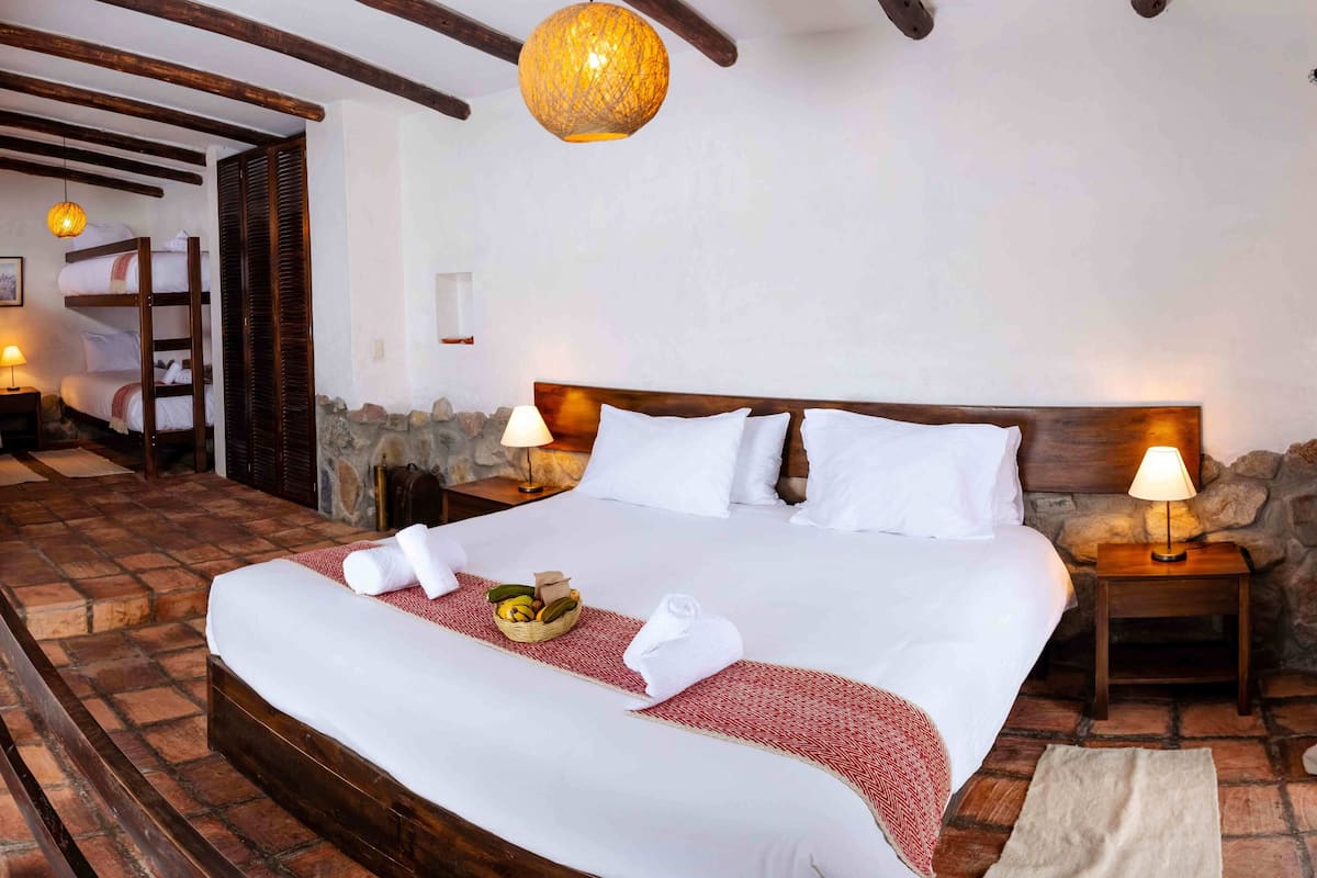 A neatly made bed with white linens, a red and white patterned throw, and a basket of fruit on the bed. There are two bedside lamps, a wooden headboard, and a stone wall behind the bed.
