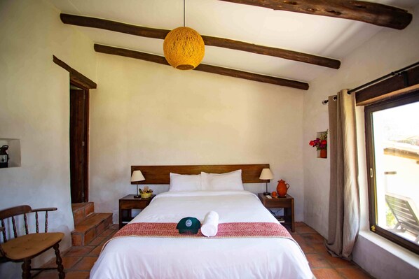 Une chambre d’hôtel avec une tête de lit en bois, un lit recouvert de draps blancs, une couvre-lit à rayures rouges et blanches, une casquette verte et une serviette enroulée posée sur le lit. On y trouve également une chaise en bois, une table de chevet avec une lampe et une fenêtre avec des rideaux.