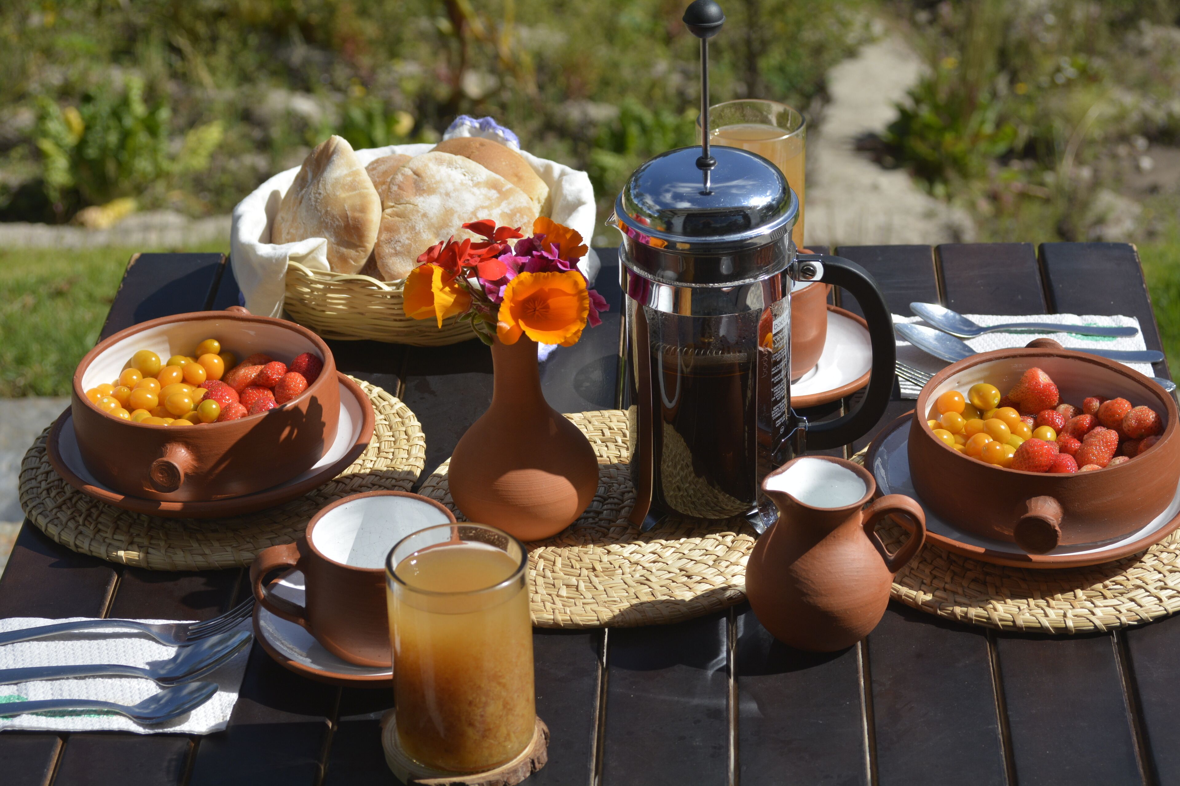 A table set with a coffee press, bowls of fruit, a basket of bread, and a pitcher of milk.