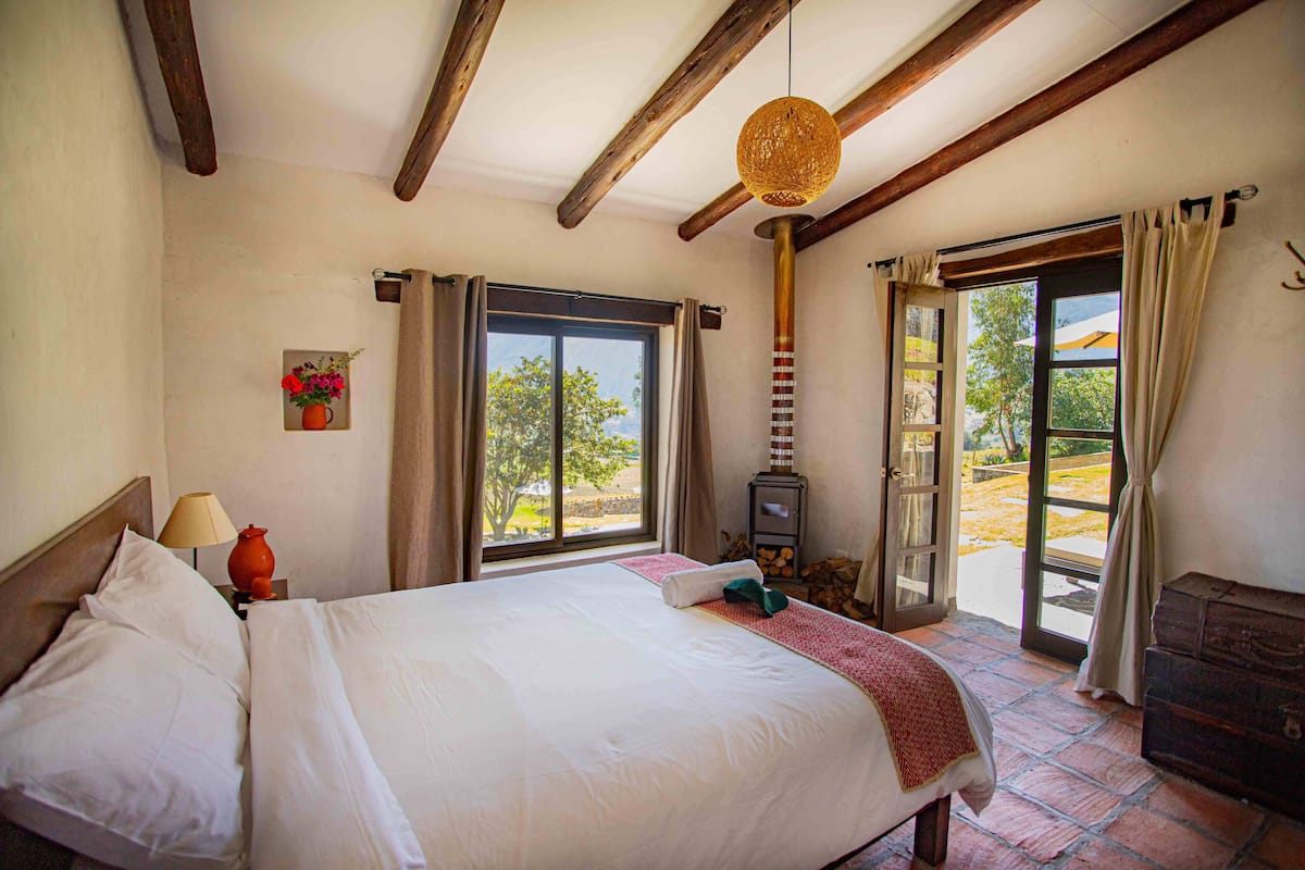 A bedroom with a bed, a nightstand, a lamp, a vase with flowers, a window, and a door leading to an outdoor area.