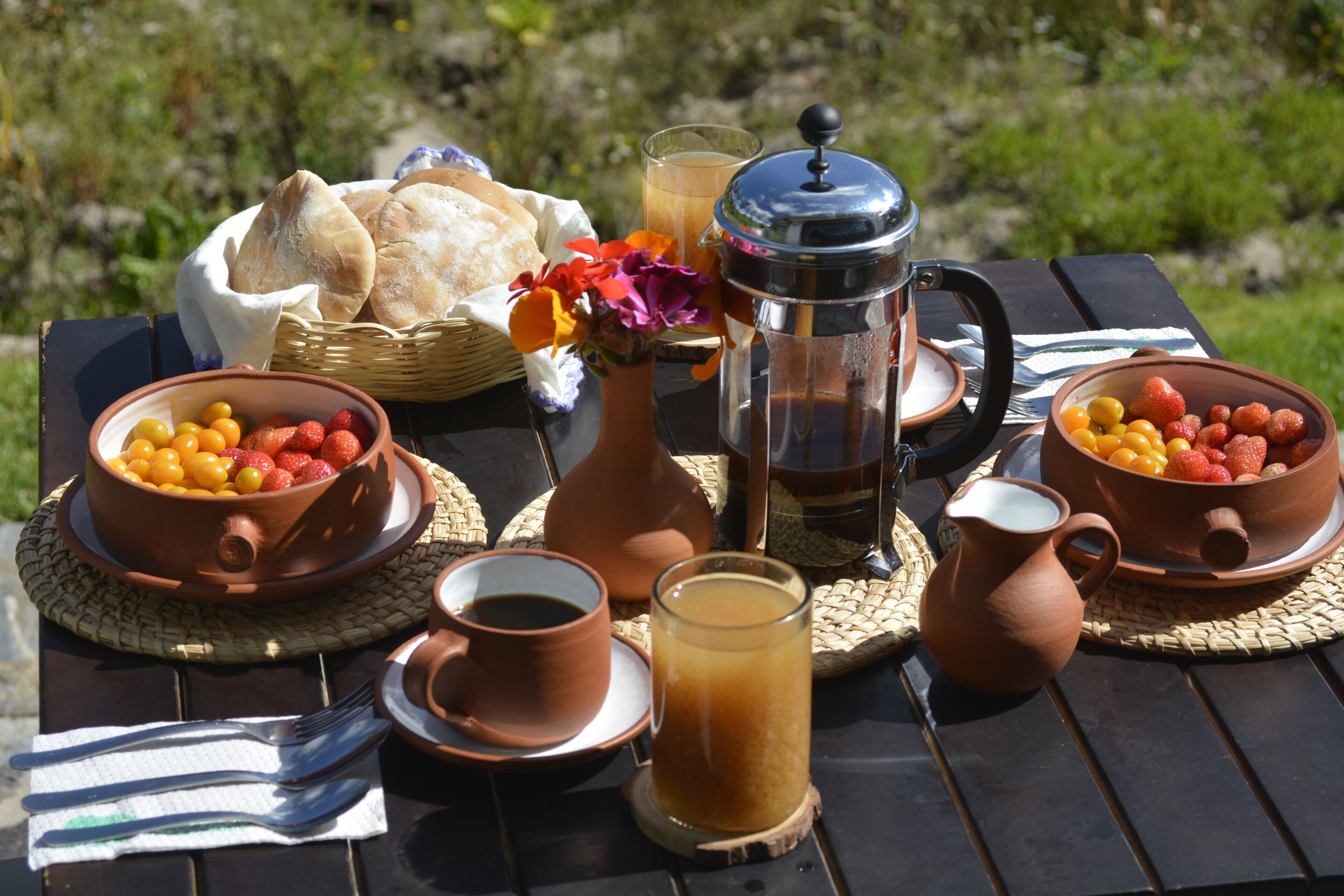 Ein Picknick mit einem Korb voller Brot, Schüsseln mit Obst, einer Kaffeemaschine, Tassen und einem Saftkrug auf einem Tisch im Freien.