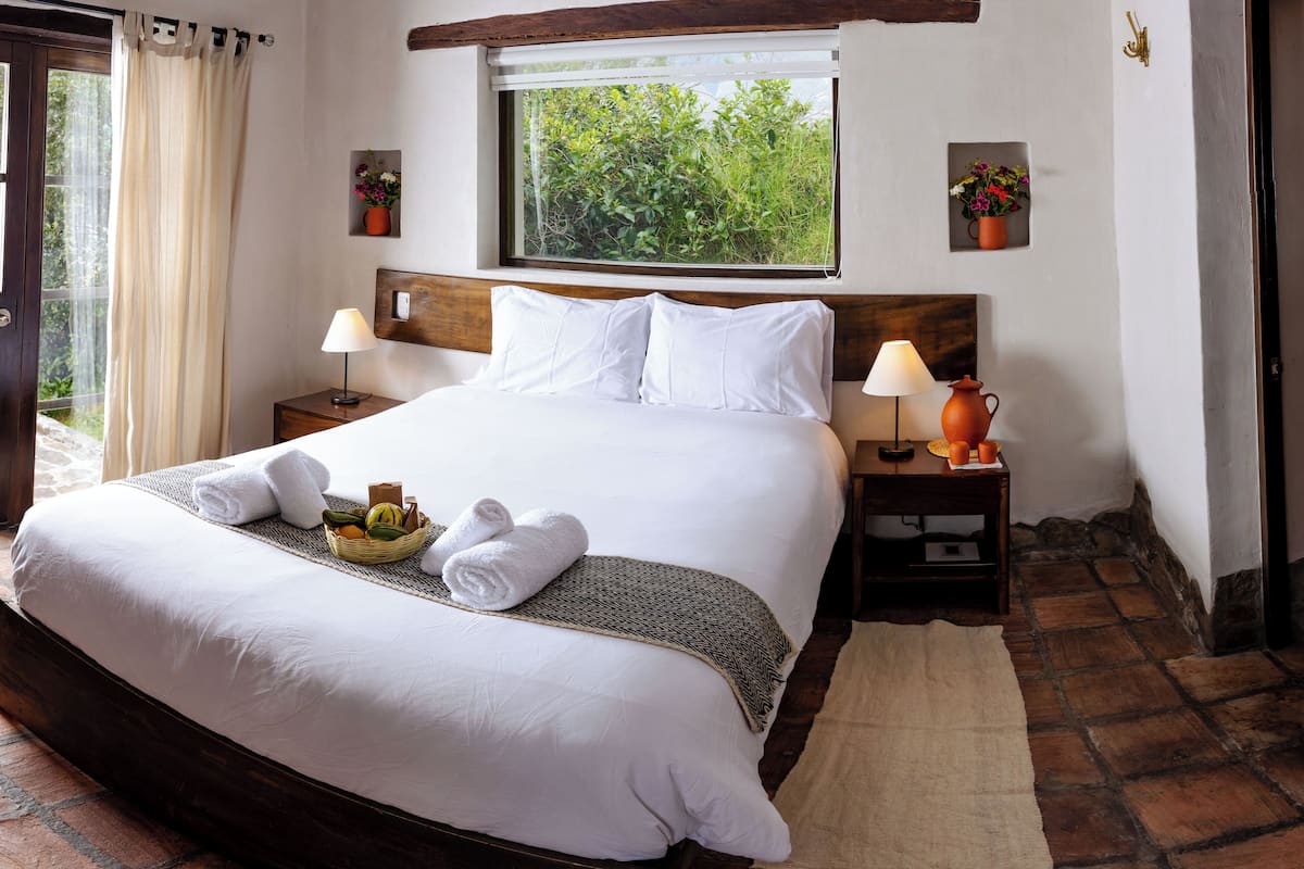 A neatly made bed with white linens, a bedside table with a lamp, and a window with a view of greenery.