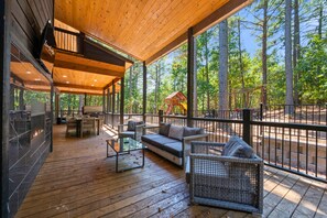 A covered outdoor deck with a sofa, chairs, and a table, overlooking a forested area. - Luxury Group Retreat with Game Room and Hot Tub (Broken Bow)