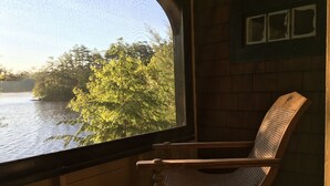 A wooden chair by a window overlooking a lake with trees.
