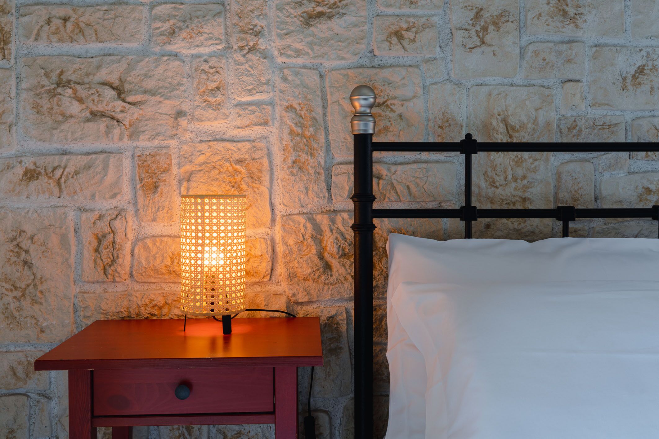 A bed with a black metal headboard, a wooden nightstand with a lamp, and a stone wall background.