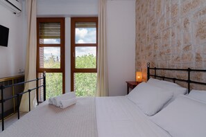 A hotel room with a bed, a nightstand, a window with blinds, and a view of greenery outside. - B&B Elegance (Bologna)