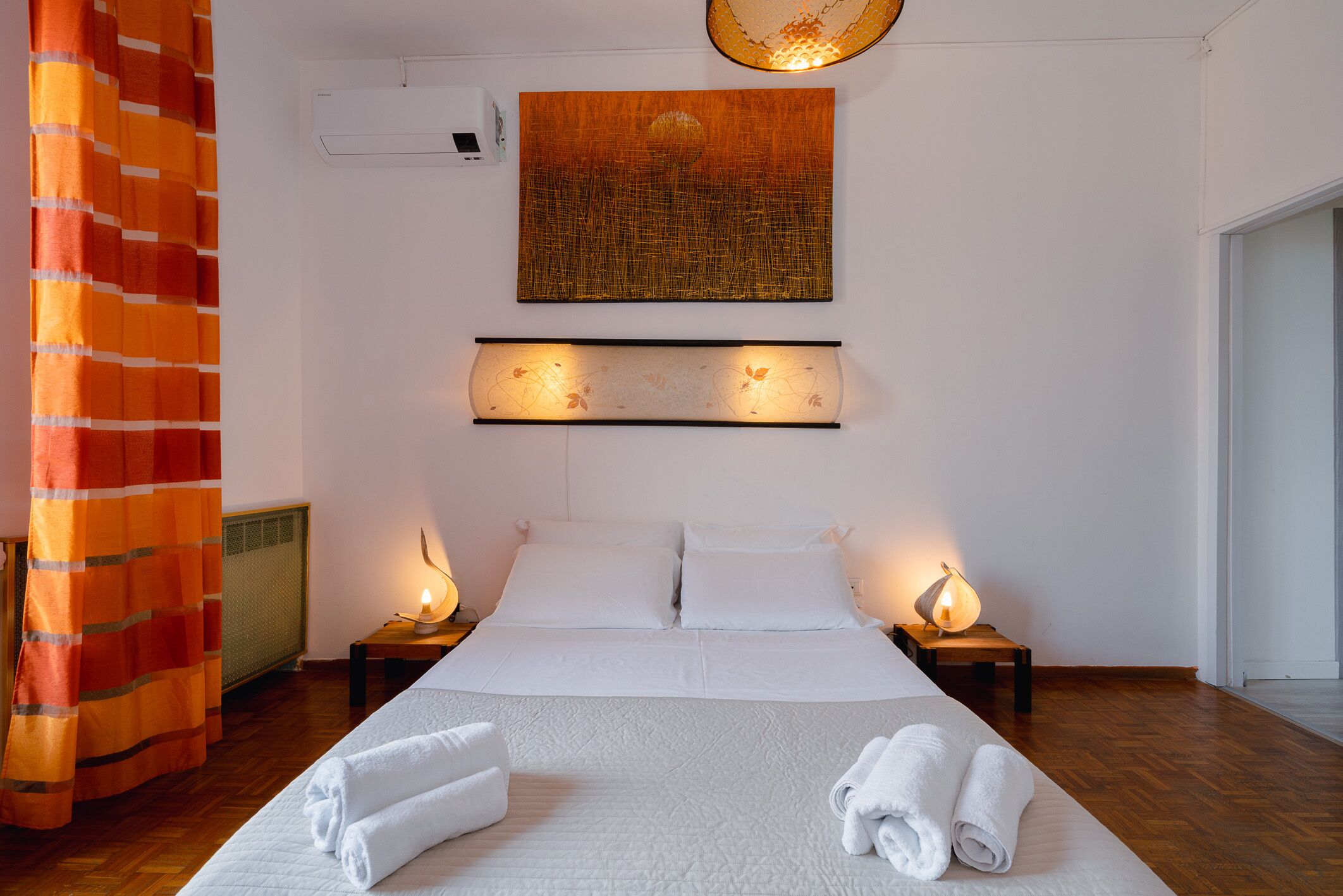 Ein Hotelzimmer mit einem großen Bett, zwei Nachttischen inklusive Lampen, einer Wand-Klimaanlage und einem großen, abstrakten Gemälde über dem Bett.