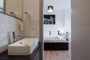 A modern bathroom with a white sink, a hairdryer, and a mirror. The room features a wooden floor and a bed with white bedding. - B&B Elegance (Bologna)